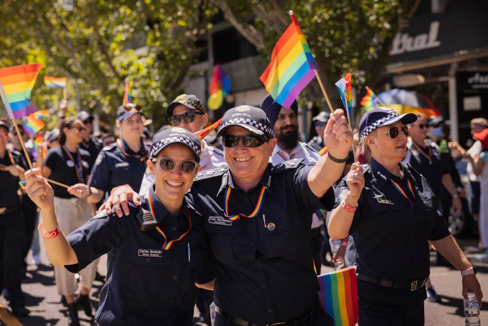 Police officers walking along the road and footpath during the day in front of shops taking part in a peaceful pride march for the LGBTQIA+ community. Two police officers in uniform are smiling at the camera waving rainbow-coloured flags waving 