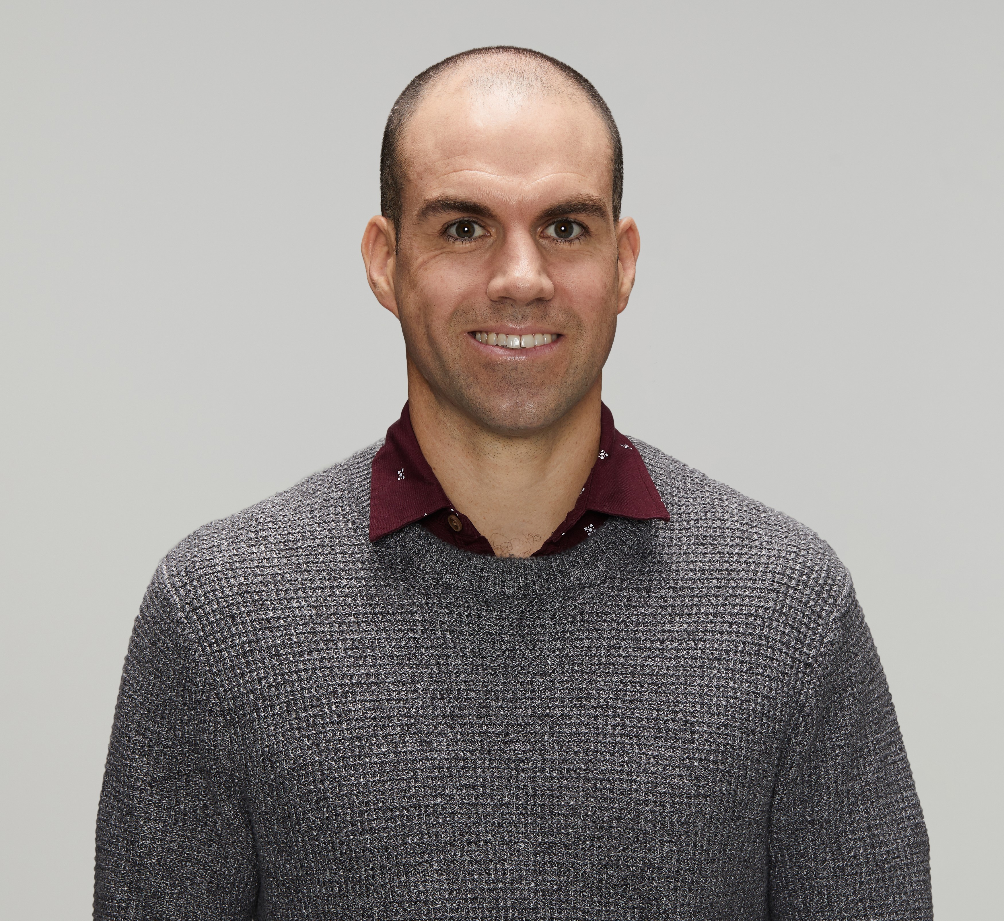 Police officer in civilian clothing smiling at the camera. White male, early forties with dark receding hair, wearing a maroon collared shirt with a smart casual metal grey jumper over the top.