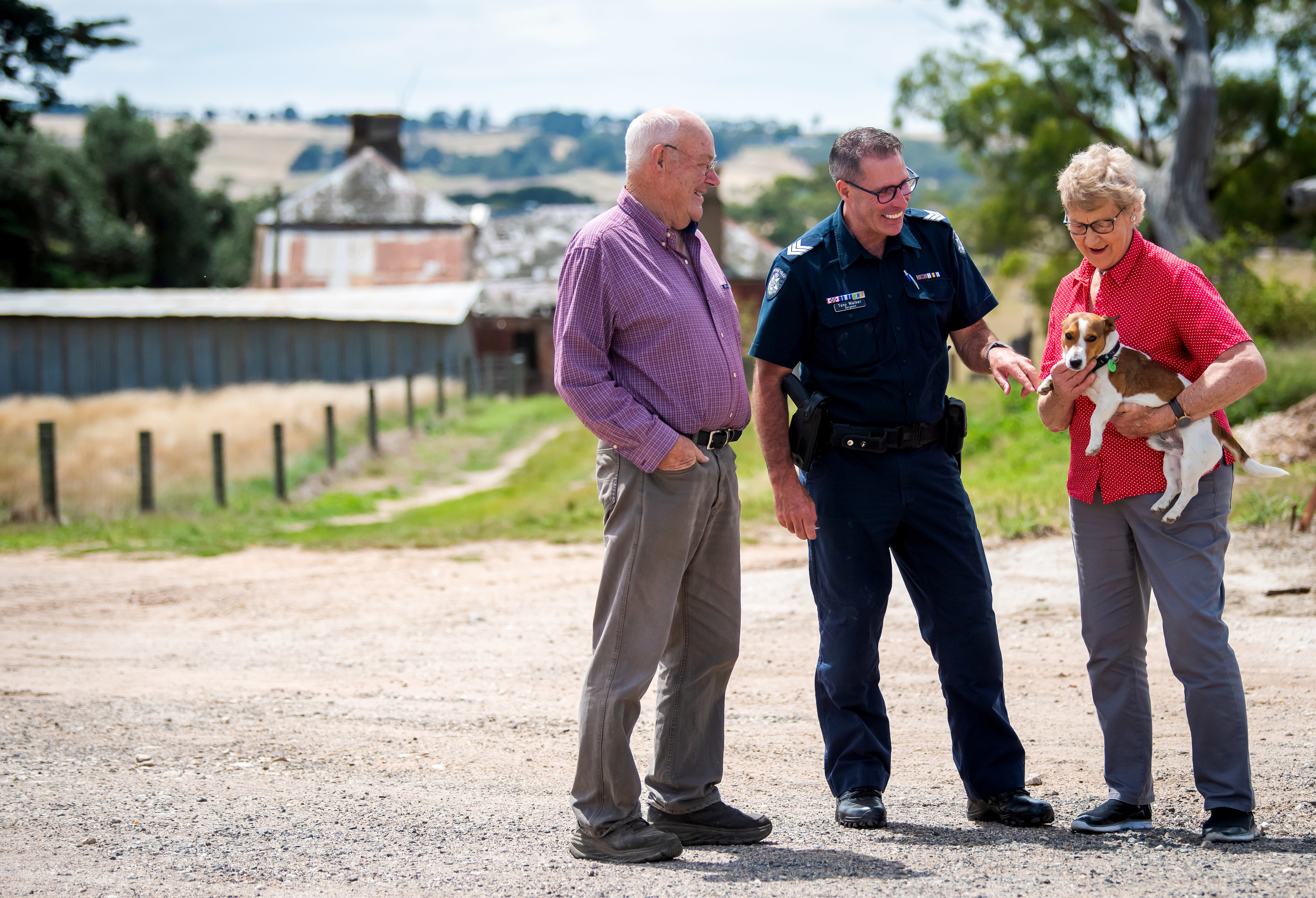 A uniformed police officer smiling and chatting with an elderly male and female and their dog. They are in a rural area surrounded by farmland and trees.